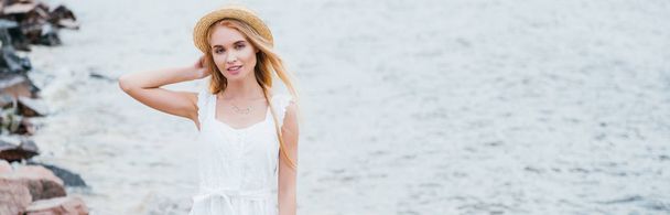 panoramic shot of cheerful young blonde woman touching straw hat and looking at camera near sea  - Фото, изображение