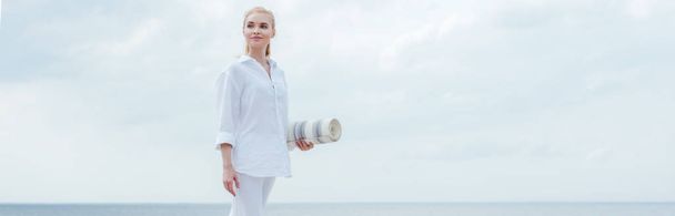 panoramic shot of happy young blonde woman standing near sea and holding yoga mat  - Photo, image
