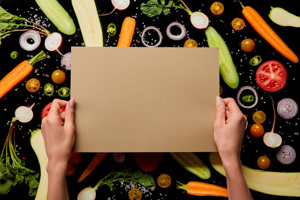 cropped view of woman holding empty paper on vegetable pattern background isolated on black - Foto, Imagem