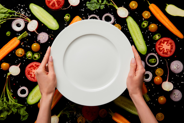 cropped view of woman holding empty round plate on vegetable pattern background isolated on black - Foto, afbeelding