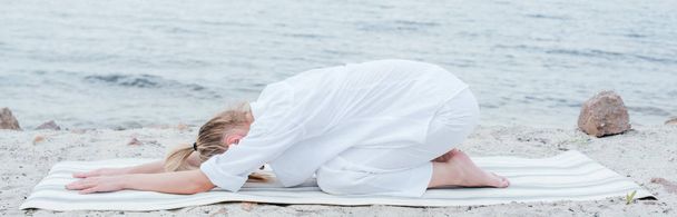plano panorámico de mujer joven rubia relajándose y practicando yoga en estera de yoga cerca del mar
  - Foto, Imagen