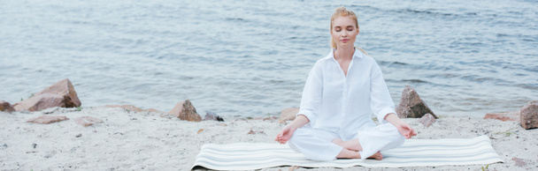 Panoramaaufnahme einer friedlichen blonden jungen Frau mit geschlossenen Augen, die in Flussnähe Yoga praktiziert  - Foto, Bild