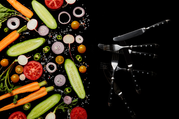 top view of fresh vegetable slices with salt near knives and forks isolated on black with copy space - 写真・画像