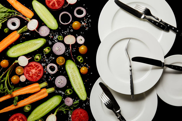 top view of round plates and cutlery near sliced vegetables isolated on black - Foto, Imagem