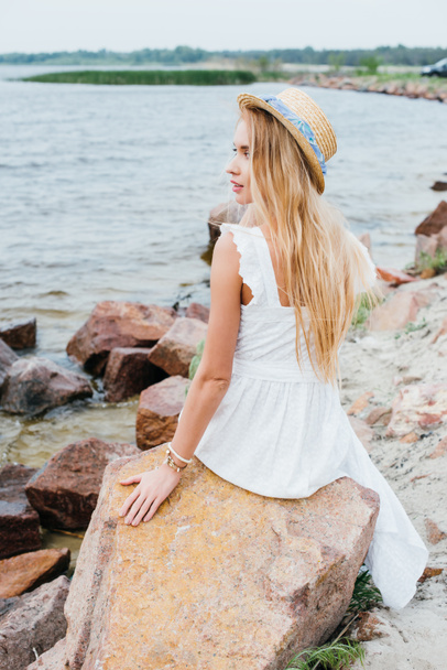 belle femme blonde en chapeau de paille assise sur des pierres près de la mer
  - Photo, image