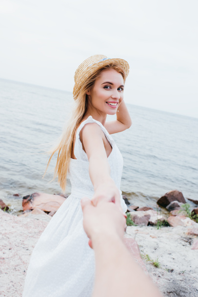 abgeschnittene Ansicht eines Mannes mit glücklicher blonder Frau im Strohhut an der Hand  - Foto, Bild