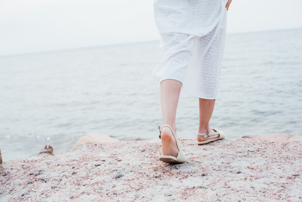 cropped view of young woman in white dress and sandals standing near sea  - Photo, image