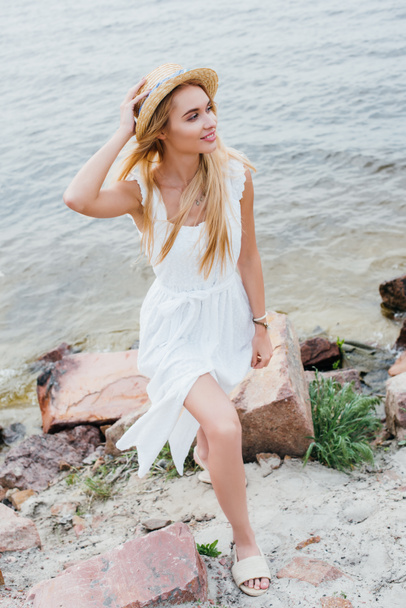 alegre joven mujer tocando sombrero de paja y sonriendo mientras está de pie cerca del mar
  - Foto, imagen