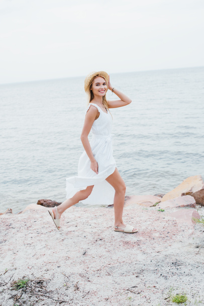 feliz chica rubia sonriendo mientras toca sombrero de paja y caminando cerca del mar
  - Foto, imagen