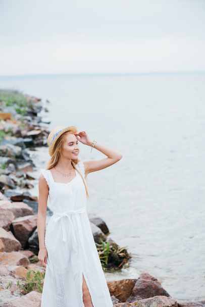 cheerful young blonde woman touching straw hat and looking at sea  - Fotografie, Obrázek