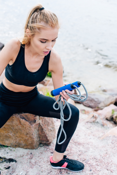 exhausted blonde young woman in sportswear holding jump rope near sea - Foto, Imagem