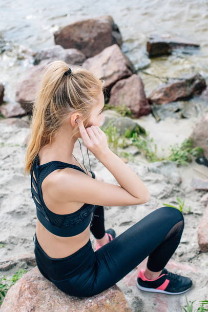 εναέρια θέα της αθλητικής γυναίκας που κάθεται στην πέτρα ακούγοντας μουσική σε ακουστικά - Φωτογραφία, εικόνα