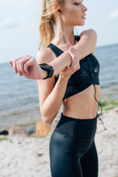 selective focus of blonde athletic girl stretching and listening music near sea  - Fotó, kép