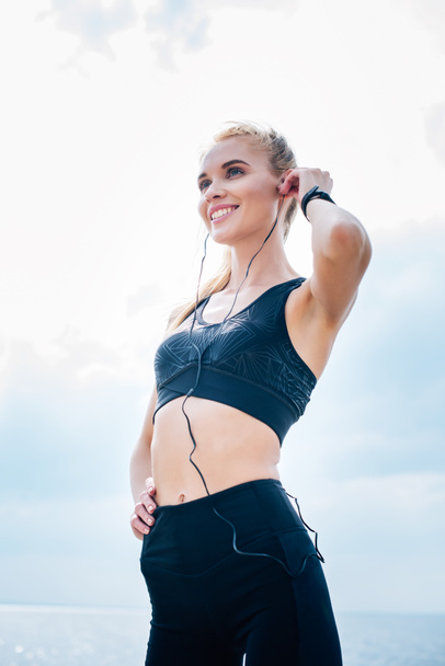 low angle view of happy athletic girl listening music while standing with hand on hip  - Фото, зображення