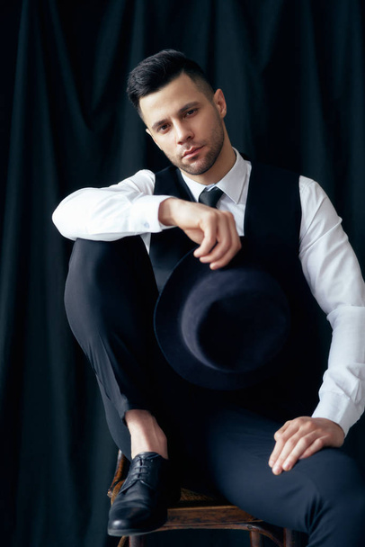 Handsome young man in elegant suit with hat in hands posing in studio on black background. Retro style - Fotó, kép