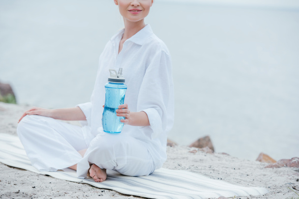 vista cortada da mulher alegre que senta-se no tapete do ioga e que prende a garrafa do esporte perto do mar
  - Foto, Imagem