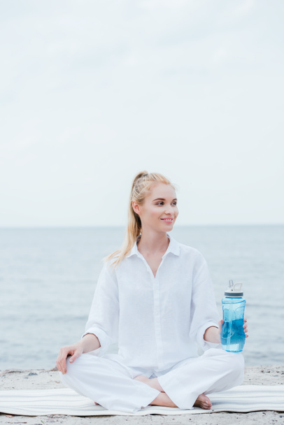 cheerful blonde woman holding sport bottle and sitting on yoga mat near sea  - Foto, imagen