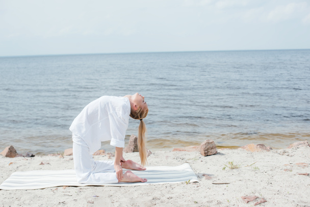 mujer bonita con los ojos cerrados practicando yoga en la estera del yoga cerca del mar
  - Foto, Imagen