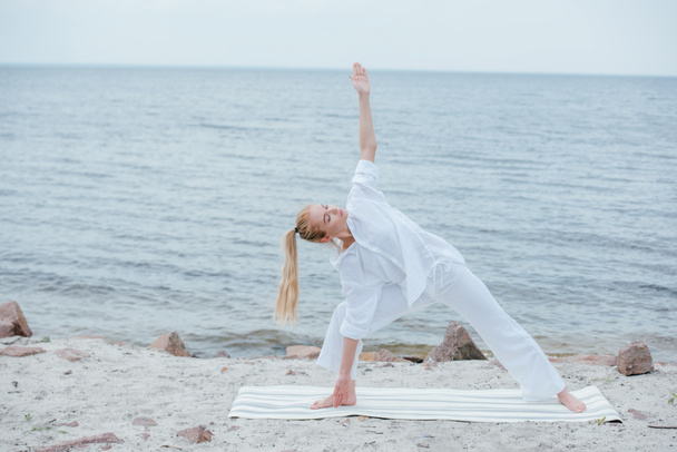 attractive girl with closed eyes practicing yoga on yoga mat near sea  - Fotografie, Obrázek