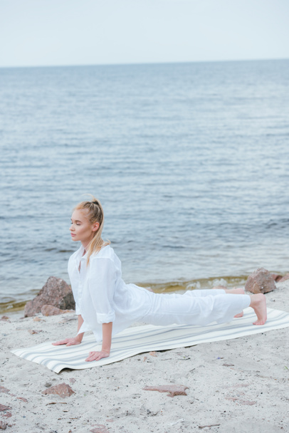 attractive blonde young woman with closed eyes practicing yoga near river on yoga mat - Foto, Imagem