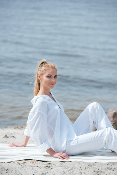 szczęśliwy i atrakcyjny blond młoda kobieta siedzi na mat jogi  - Zdjęcie, obraz
