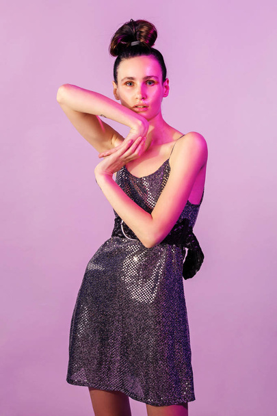 Fashion young girl posing or dancing in colorful bright neon lights. Girl in a brilliant dress hair tied in knot, gloss face in purple color light on pink background - Foto, Imagem