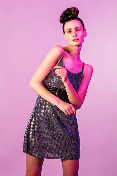 Fashion young girl posing or dancing in colorful bright neon lights. Girl in a brilliant dress hair tied in knot, gloss face in purple color light on pink background - Φωτογραφία, εικόνα