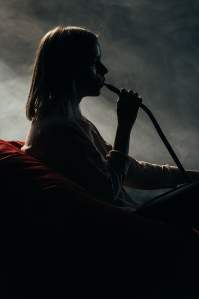 silhouette of woman sitting on bean bag chair and smoking hookah in darkness - Photo, Image