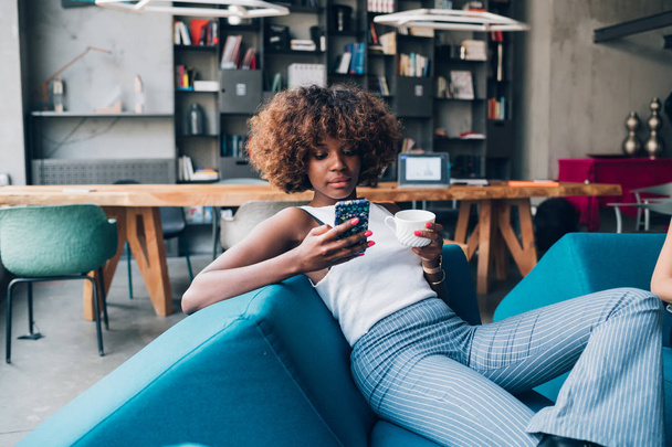 young black woman drinking and chatting with smartphone in modern loft - Foto, imagen