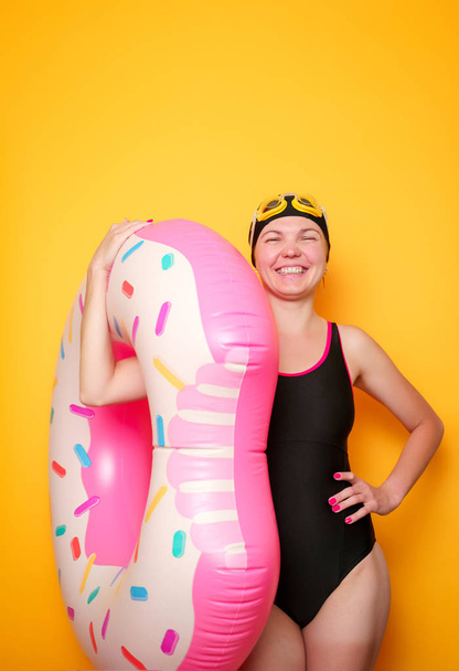 Image of girl in swimming goggles, black swimsuit with donut lifebuoy on empty orange background - Foto, afbeelding