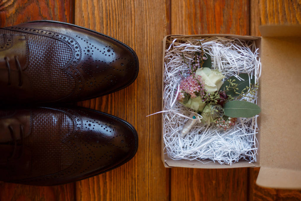 Brown men's boots stand on a wooden plank floor next to a cardboard box. In the package is a filler of decorative straw and the groom's boutonniere of small lilac flowers, white roses and green leaves. - Valokuva, kuva