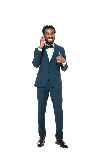 hombre afroamericano feliz en traje hablando en el teléfono inteligente y mostrando el pulgar hacia arriba aislado en blanco
  - Foto, Imagen
