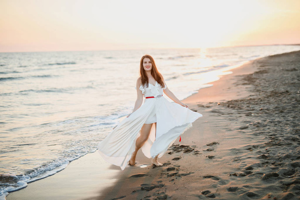 young beautiful girl in a white dress walks on the beach near the sea at sunset - Фото, зображення