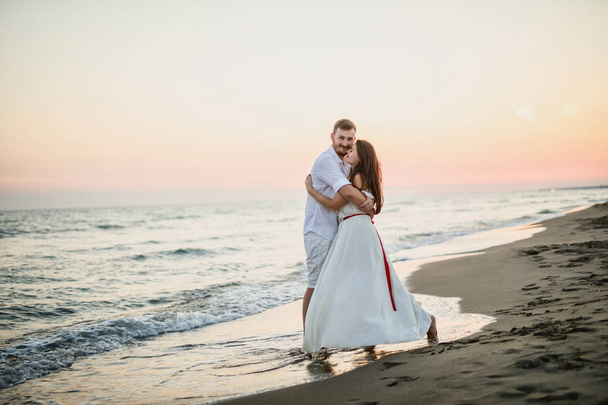 young beautiful couple in white clothes walking on the beach near the sea at sunset - Фото, изображение