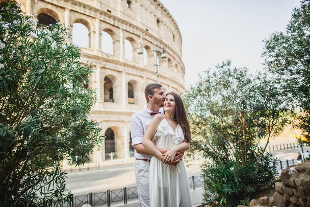 young beautiful couple in white clothes stands against the background of the Colosseum in Rome in Italy - 写真・画像