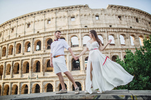 young beautiful couple in white clothes stands against the background of the Colosseum in Rome in Italy - Zdjęcie, obraz