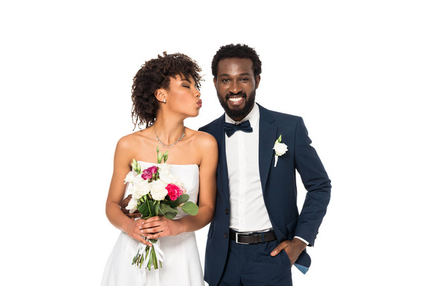 gelukkige Afro-Amerikaanse bruid met eend gezicht Holding Bouquet in de buurt van bruidegom met hand in Pocket geïsoleerd op wit  - Foto, afbeelding