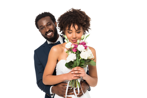 smiling african american bridegroom hugging attractive bride smelling flowers isolated on white - Foto, Bild