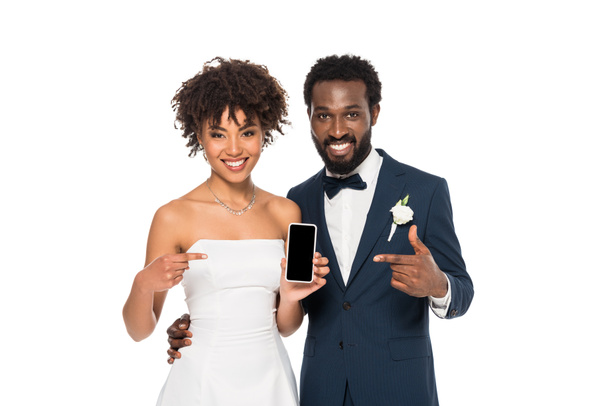 heureux marié afro-américain et mariée pointant avec les doigts sur smartphone avec écran blanc isolé sur blanc
  - Photo, image