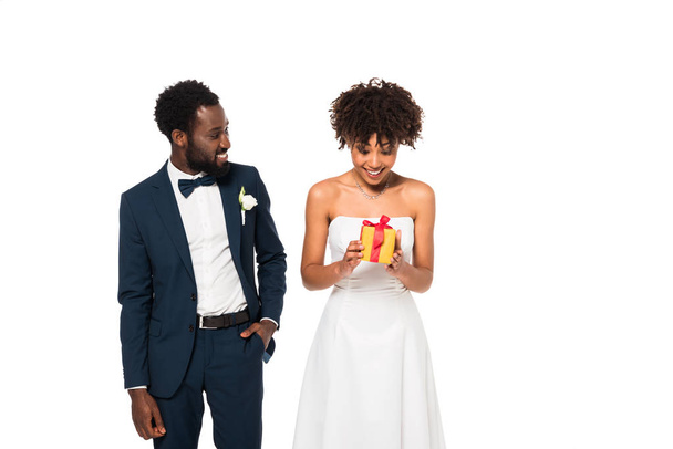 ευτυχισμένος Αφρικανός Αμερικανός γαμπρός στέκεται με το χέρι στην τσέπη και κοιτάζοντας χαρούμενα νύφη κρατώντας κουτί δώρου απομονωμένο σε λευκό  - Φωτογραφία, εικόνα