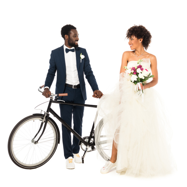 pohledný africký americký ženich poblíž nevěsty s květinami, držící jízdní kolo izolované na bílém  - Fotografie, Obrázek