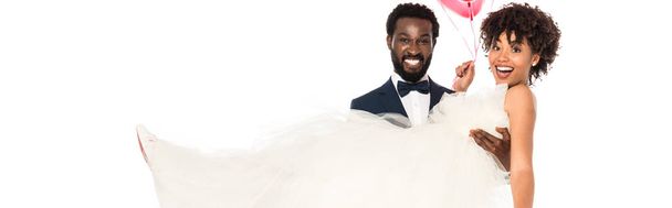 panoramic shot of handsome african american bridegroom holding in arms bride in wedding dress isolated on white  - Photo, Image