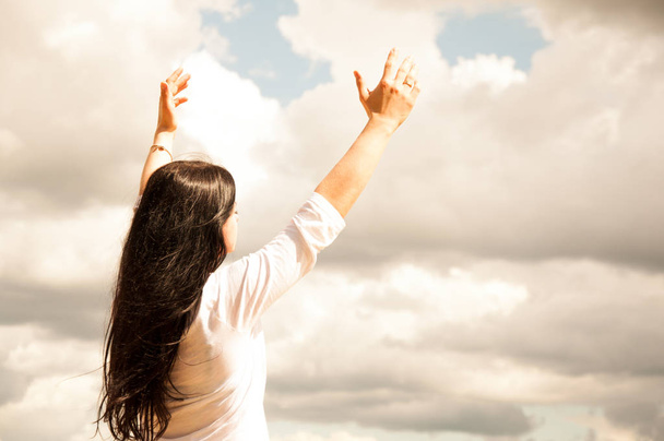 μια γυναίκα στέκεται με τα χέρια της σηκωμένο και προσεύχεται στο Θεό. Σε ένα πράσινο χωράφι το καλοκαίρι. Μπλε ουρανός και λευκά σύννεφα. - Φωτογραφία, εικόνα