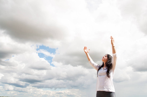 una mujer está de pie con los brazos levantados y ora a Dios. En un campo verde en verano. Cielo azul y nubes blancas
. - Foto, imagen