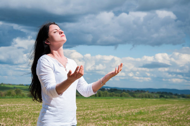 μια γυναίκα στέκεται με τα χέρια της σηκωμένο και προσεύχεται στο Θεό. Σε ένα πράσινο χωράφι το καλοκαίρι. Μπλε ουρανός και λευκά σύννεφα. - Φωτογραφία, εικόνα