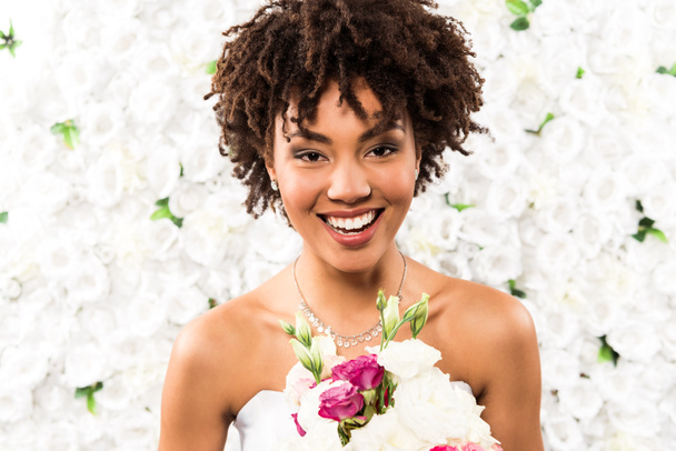 pocitive african american bride looking at camera while holding flowers  - Фото, изображение