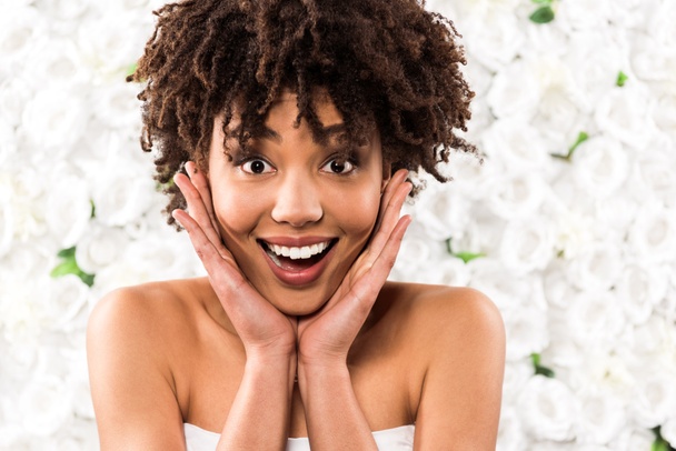 feliz noiva afro-americana tocando rosto enquanto olha para a câmera perto de flores
  - Foto, Imagem