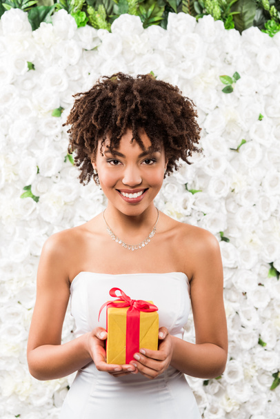 花の近くにプレゼントを保持する陽気な巻き毛アフリカ系アメリカ人の花嫁  - 写真・画像