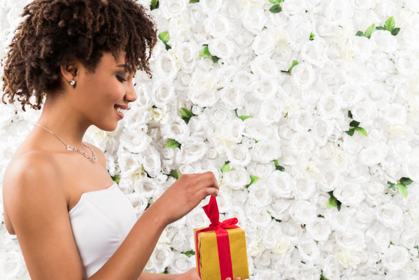 Seitenansicht der fröhlichen lockigen afrikanisch-amerikanischen Braut mit Geschenk in der Nähe von Blumen  - Foto, Bild