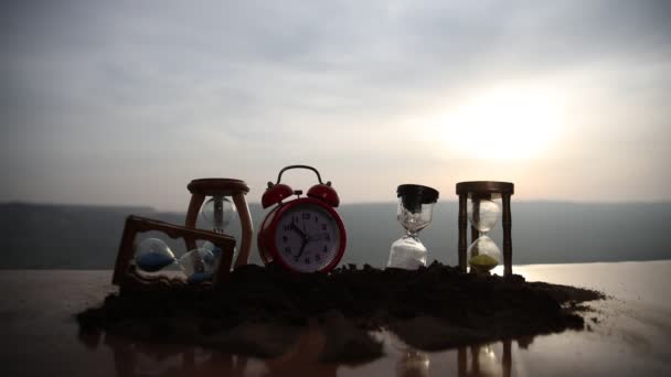 Zandloper en rode wekker tijdens zonsondergang. Tijd concept. Selectieve focus. - Video
